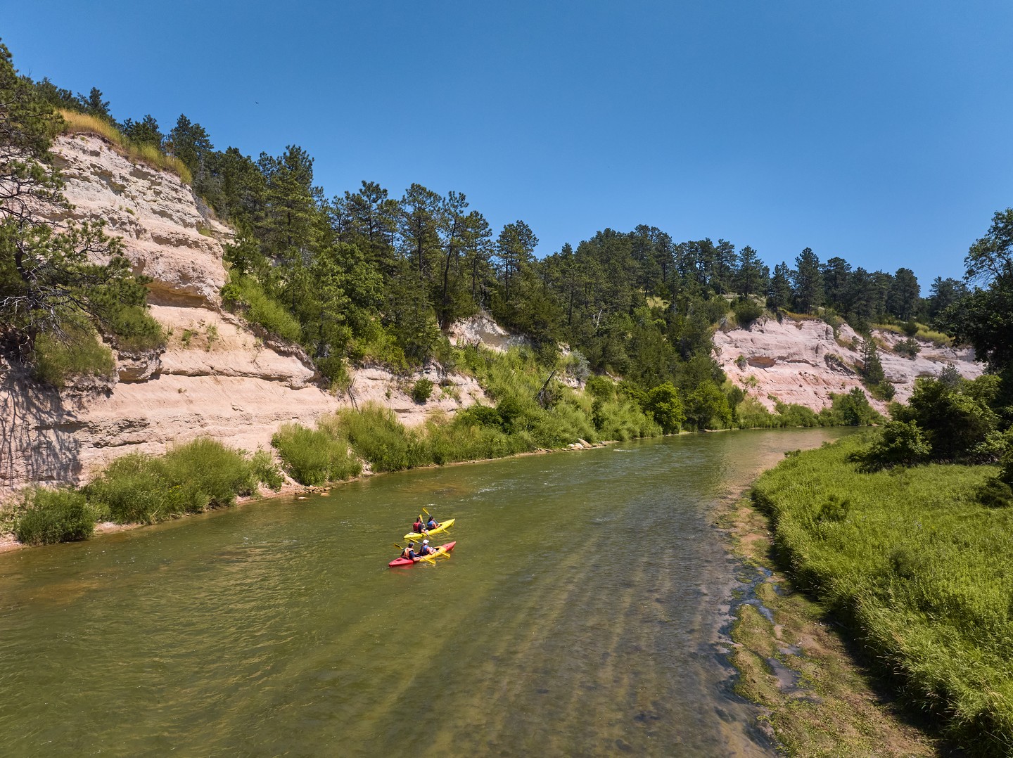 Kayakers on the Niobrara National Scenic River, with green vegetation on each side of the river and bluffs on the left – all underneath a clear blue sky. 