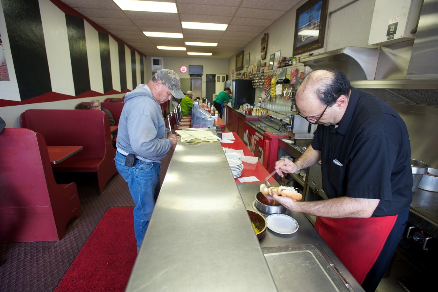 Owner George adding chili to hot dogs behind the counter with a customer standing on the other side at Coney Island Lunch Room.