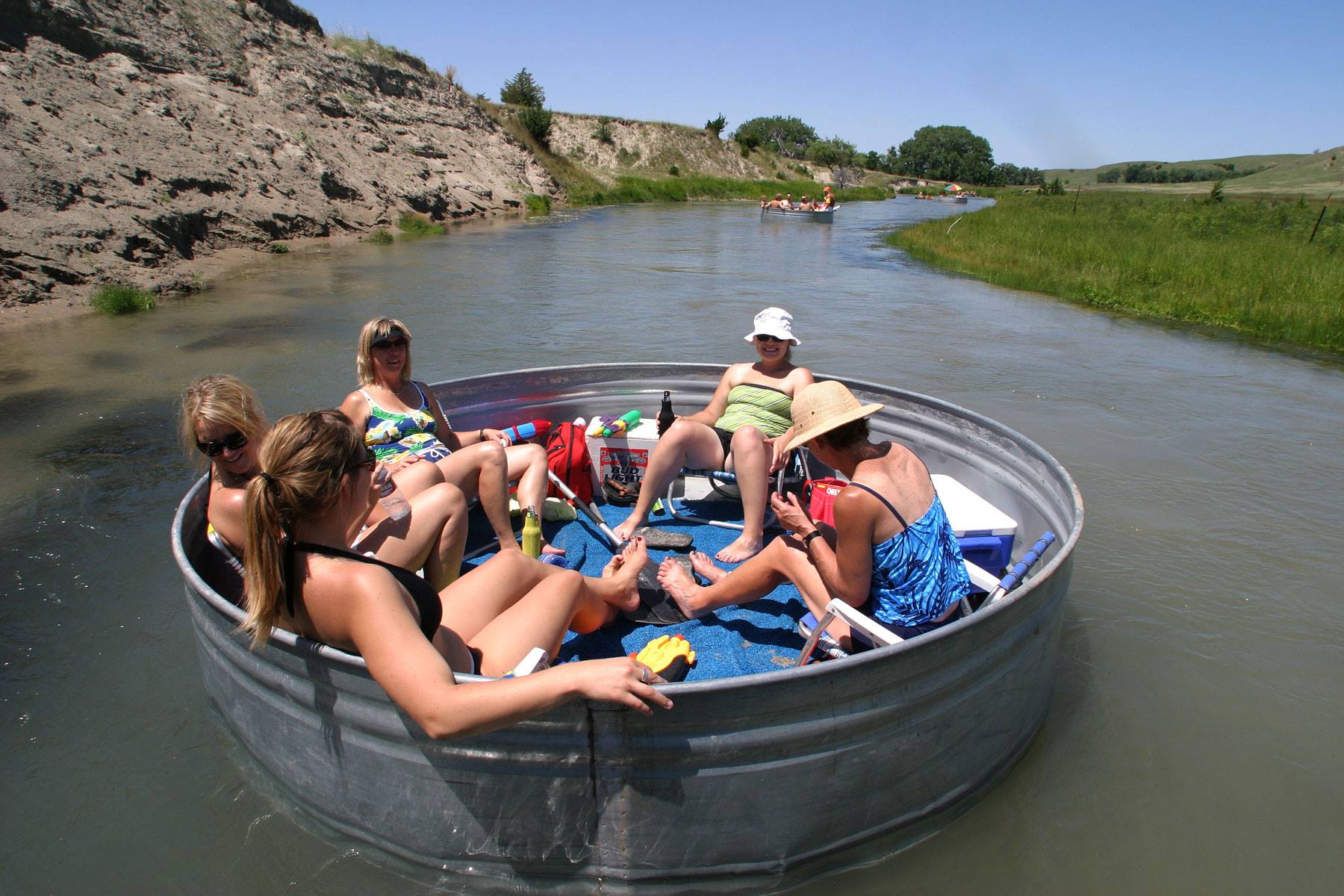A group of adults sips drinks and lounge in swimsuits in a metal drum as they float down the river with Glidden Canoe Rental near Mullen, Nebraska.