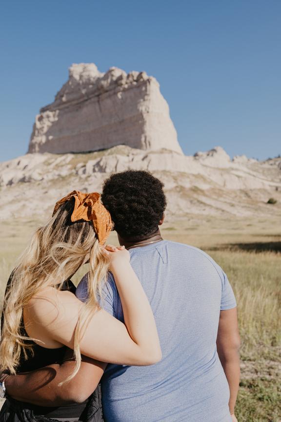 A couple stands next to a replica pioneer wagon on a trail at Scotts Bluff National Monument looking at Scotts Bluff towering in the background.