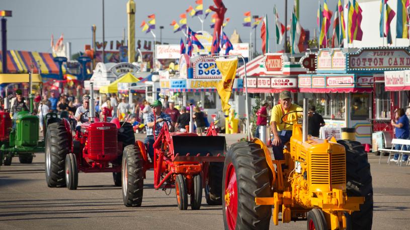 A parade of tractors riding past carnival booths at the Nebraska State Fair.